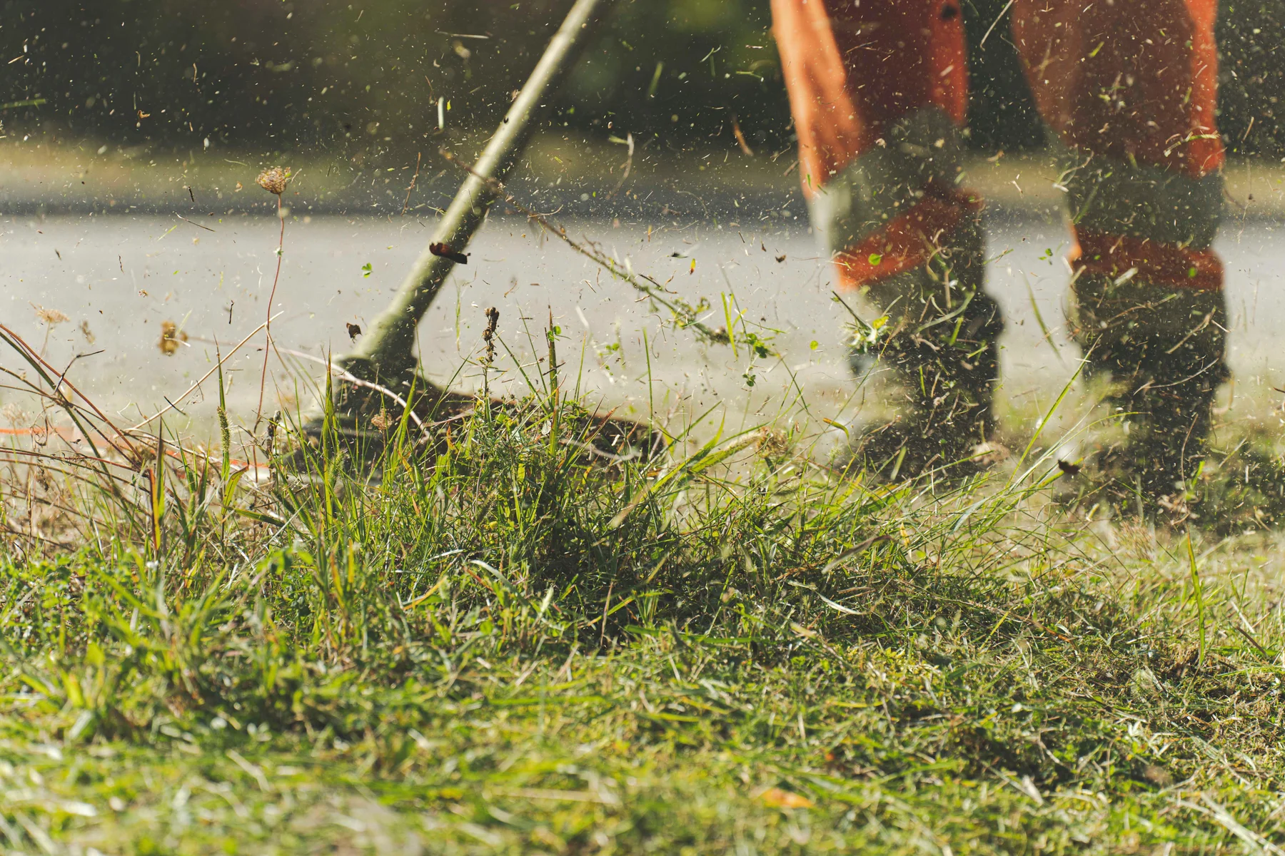 A worker in work trousers and boots uses a brush cutter to trim grass beside a path.